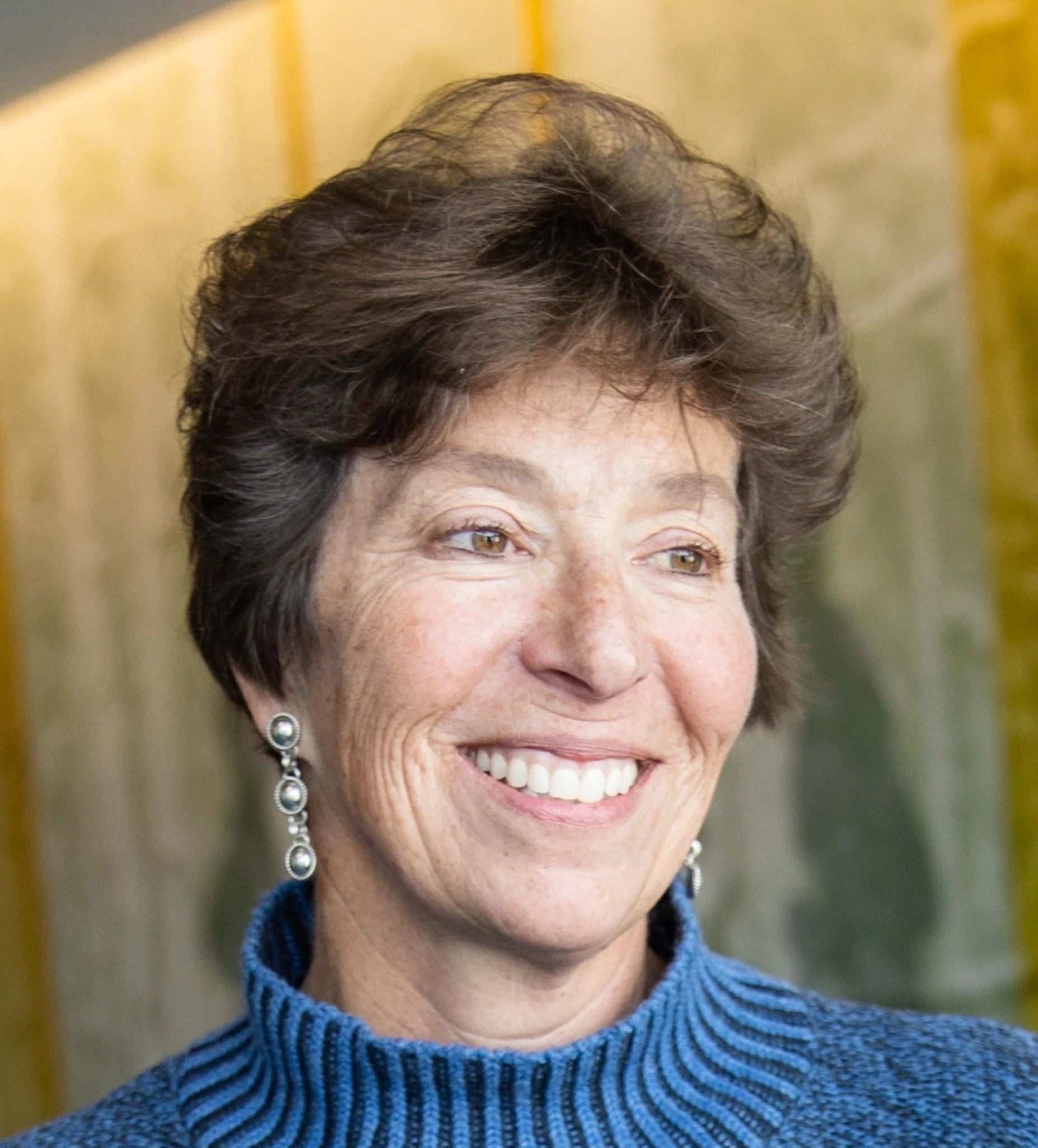 Close-up photo of a smiling middle-aged woman with short brown hair, wearing dangling earrings and a blue turtleneck sweater, with a blurred background.
