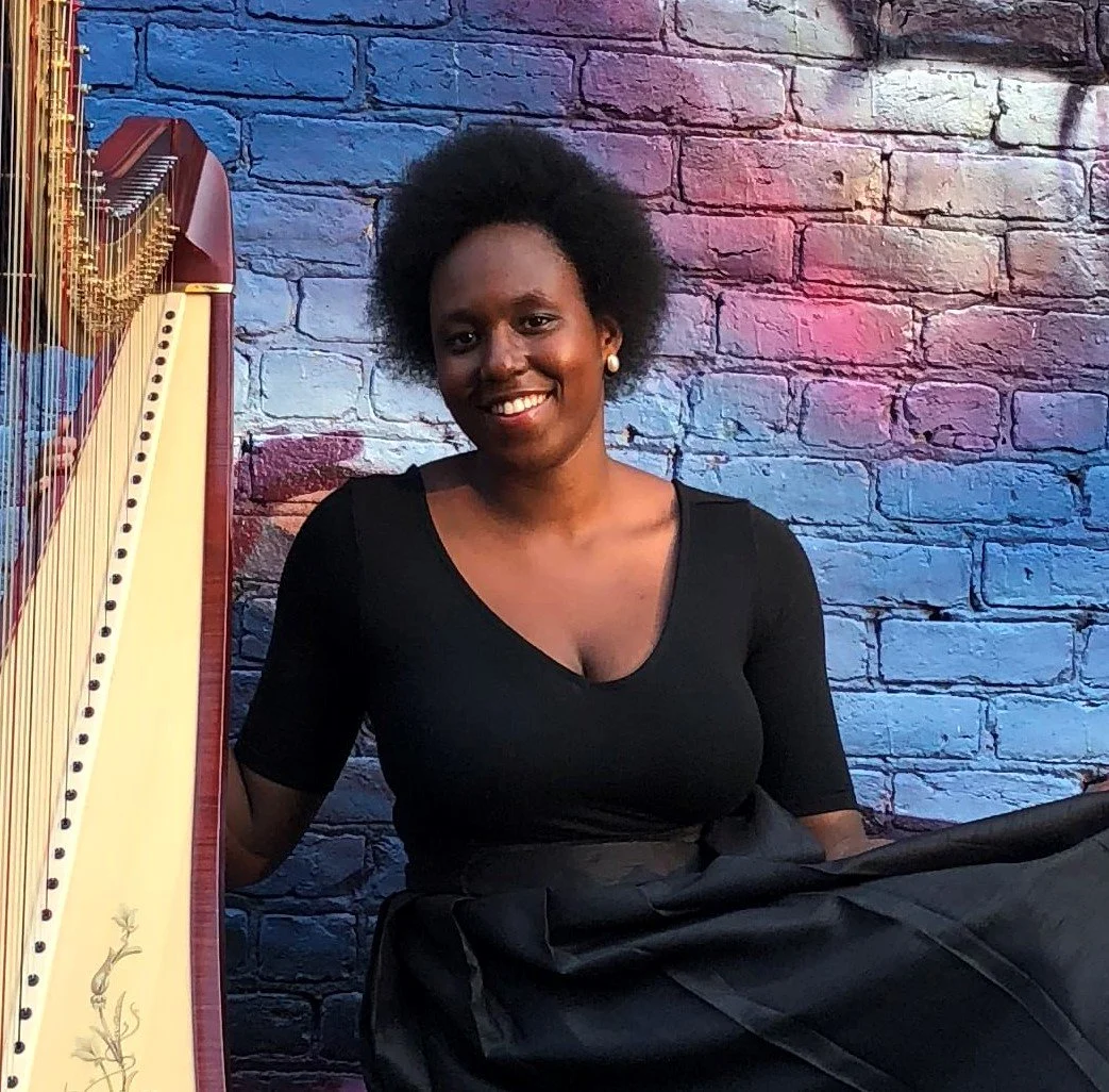 A smiling woman with curly hair, wearing a black top and earrings, sitting beside a harp against a colorful brick wall background.