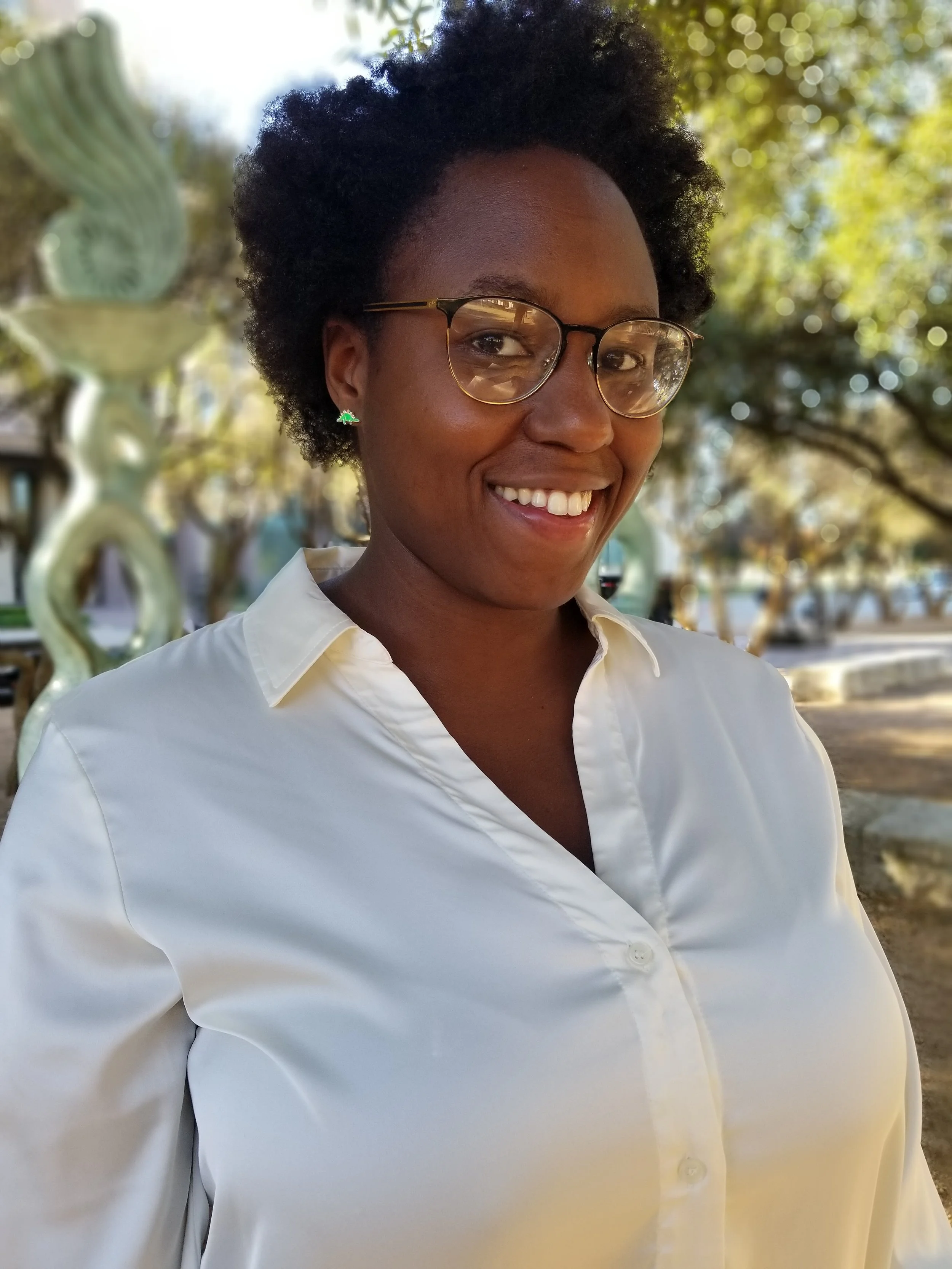 A woman with short, curly black hair and glasses smiling outdoors during daytime, wearing a white collared shirt and green Christmas tree-shaped earrings, with blurred trees and a sculpture in the background.