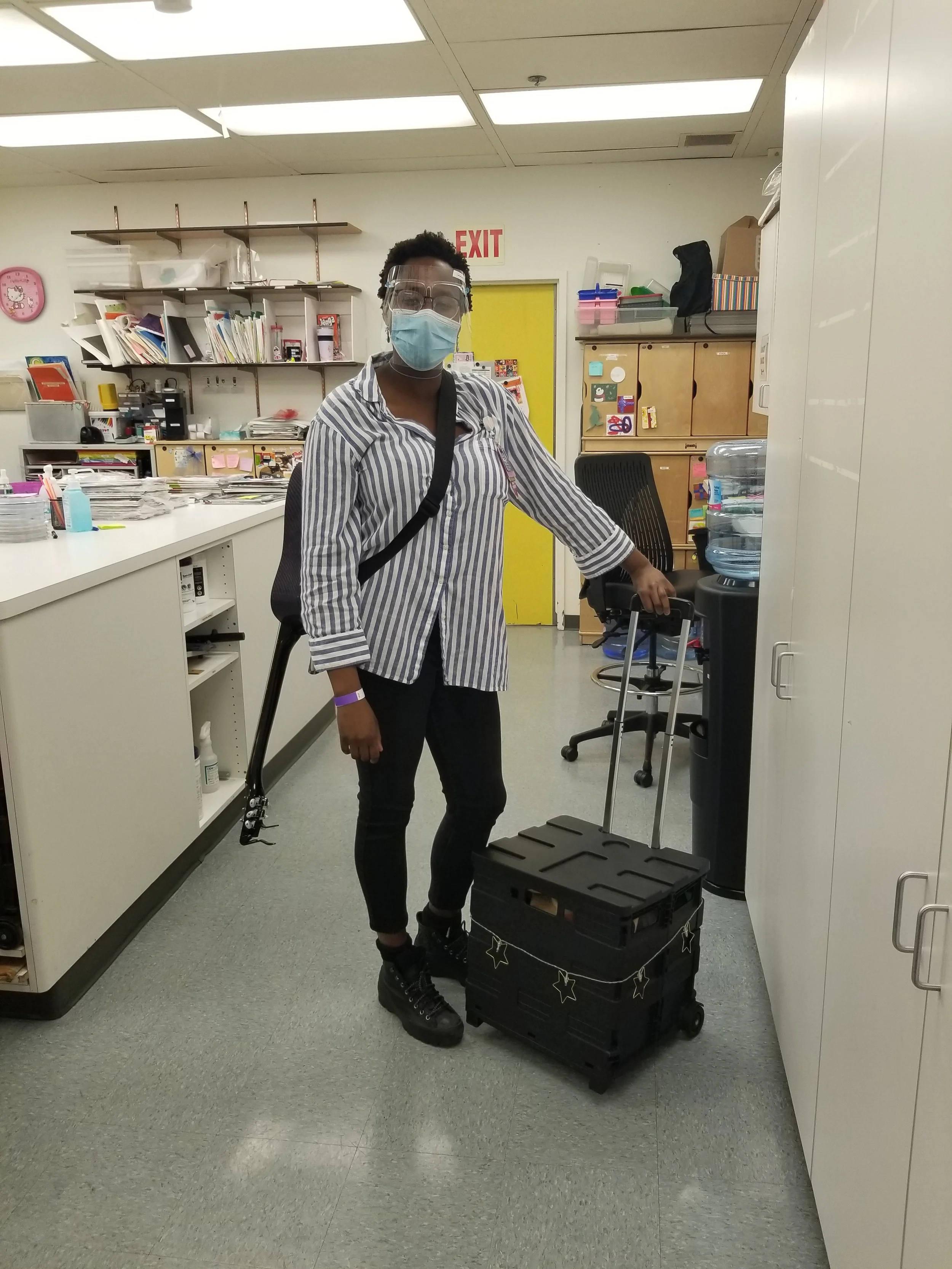 Caroline Bryan as a music therapist and expressive arts therapist at the Children's hospital of Los Angeles. She has a guitar strapped to her back, is wearing a mask and protective eye wear. She is standing by her expressive arts cart.