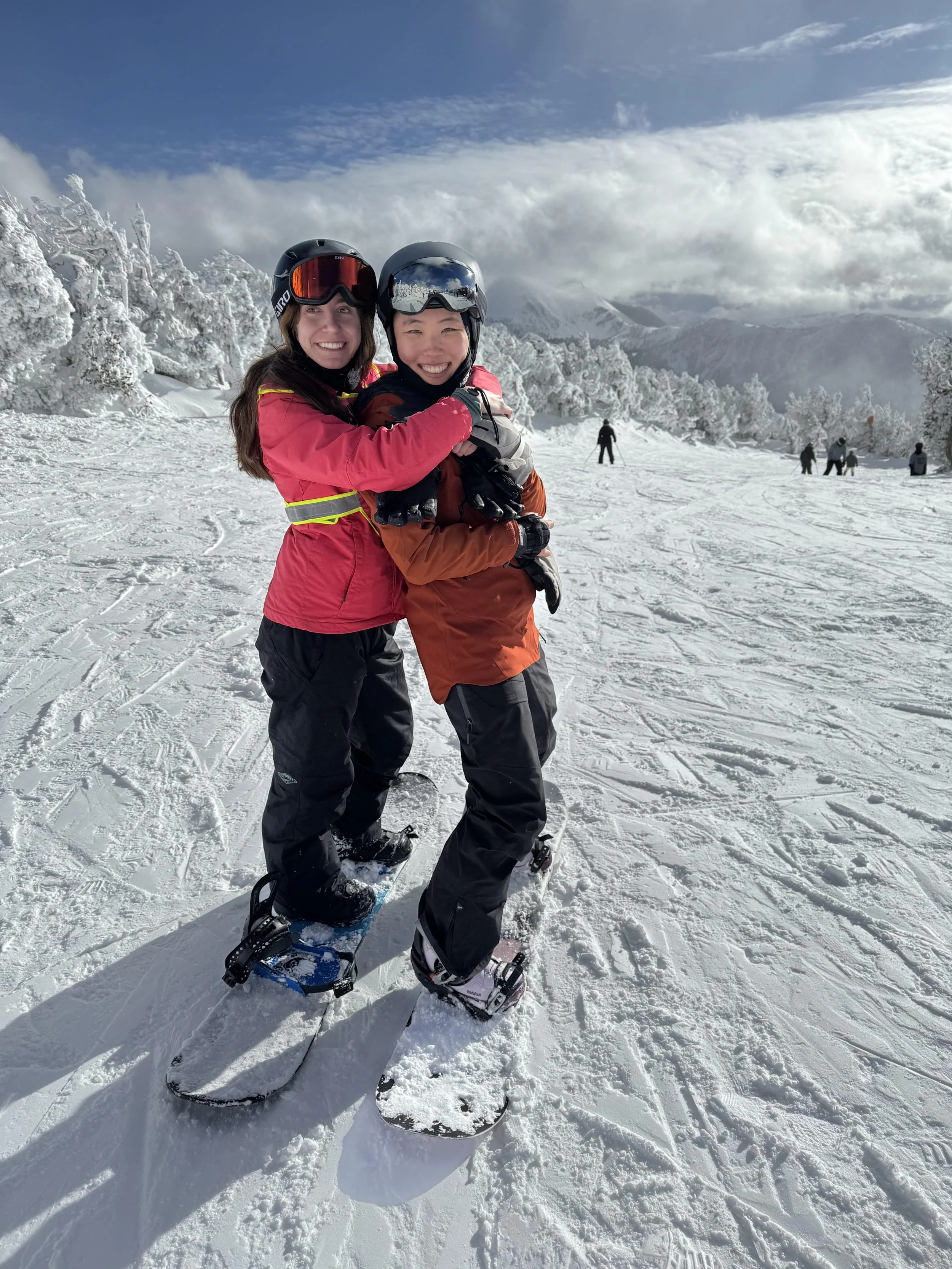 Kristie and Jungyeon smile at the camera while hugging, with each of them on their snowboard.