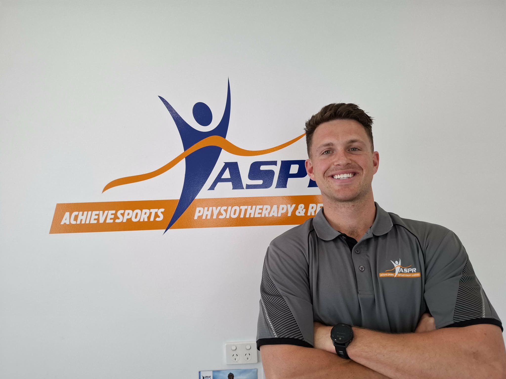 A smiling man with crossed arms standing in front of a wall with a logo and text that reads 'AchieveSports Physical Therapy & Rehab.'