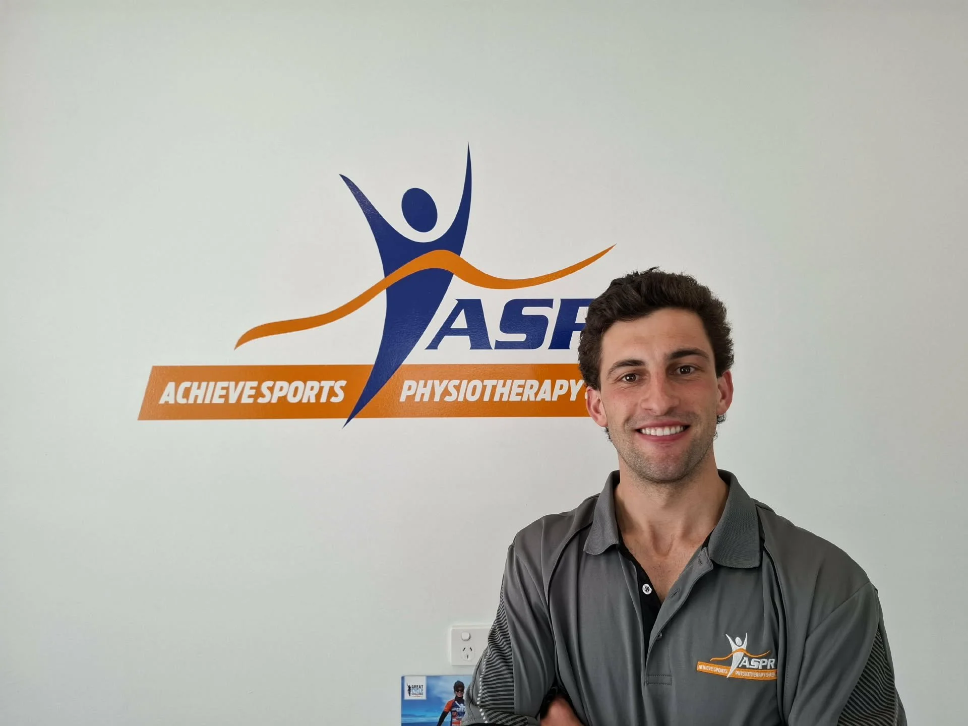 A man with brown hair, brown eyes, and olive skin, smiling, wearing a grey top with company logo, standing indoors against a plain cream-colored wall with logo in the background.