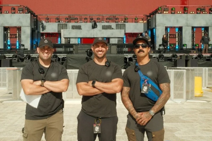 Three security personnel standing in front of a stage, smiling with arms crossed. The stage has equipment and performers in the background.