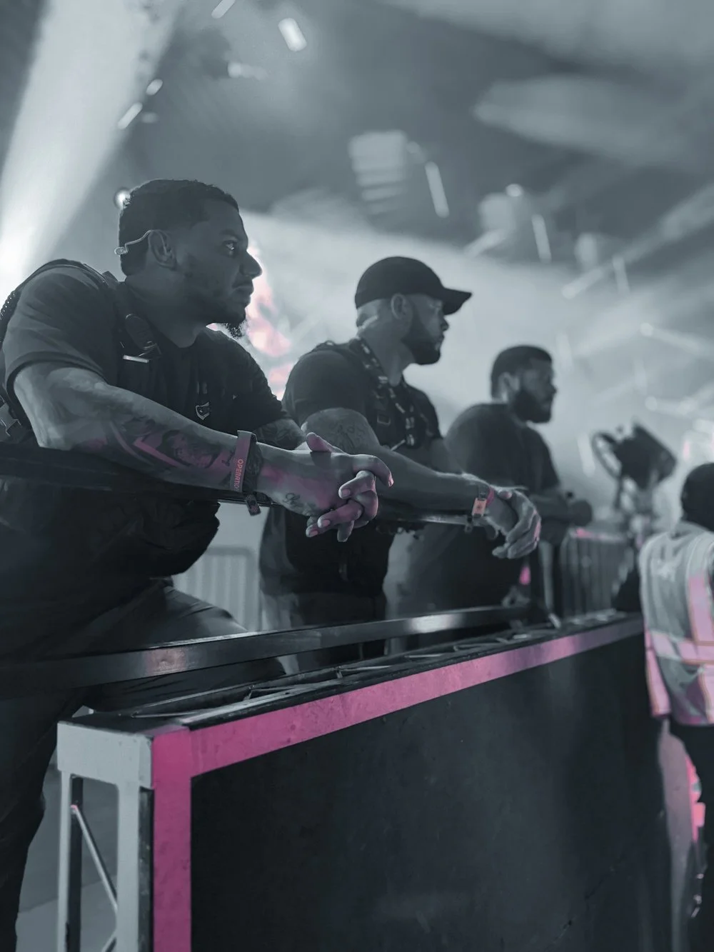 Three men with tattoos and black clothing are standing behind a barrier at a concert or event, looking towards the stage with serious expressions. The lighting is dim and atmospheric.