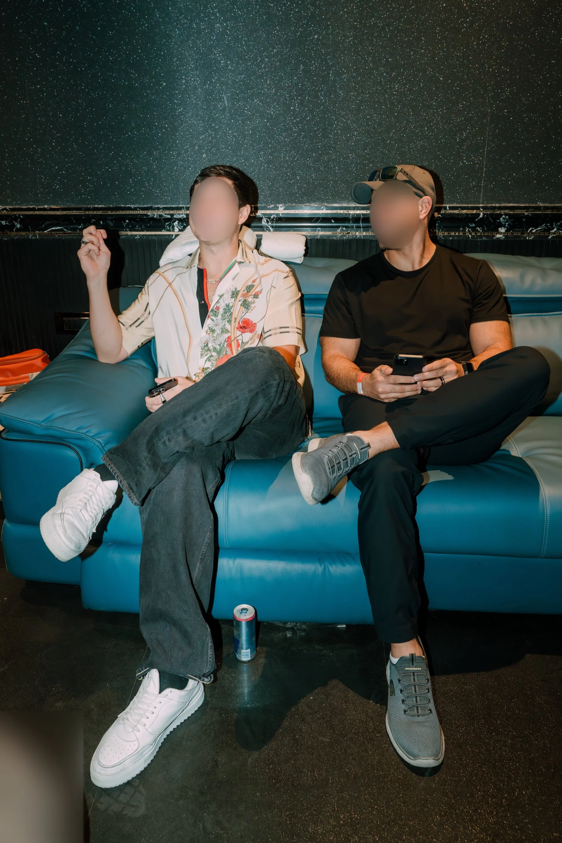 Two men sitting on a blue leather couch, one in a floral shirt and jeans, the other in a black shirt and gray sneakers, with a starry sky mural in the background.