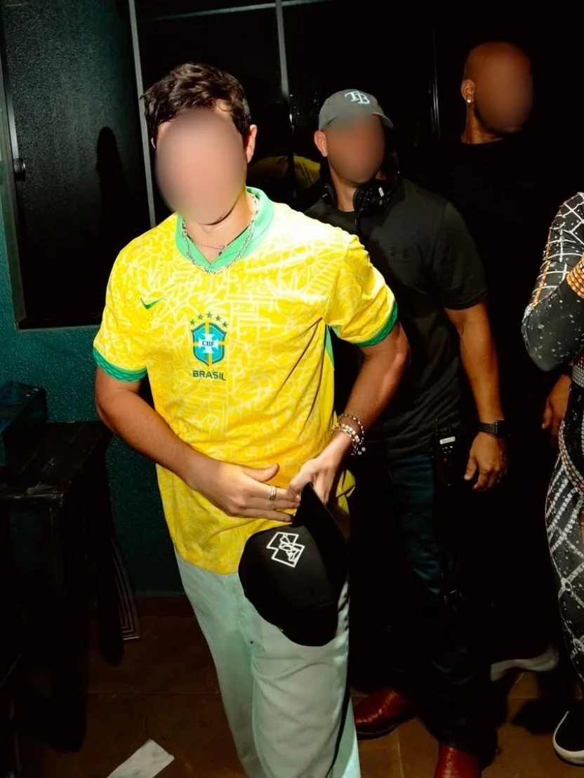 Person wearing a yellow Brazil national football team jersey holding a black cap, in an indoor setting with other people around.