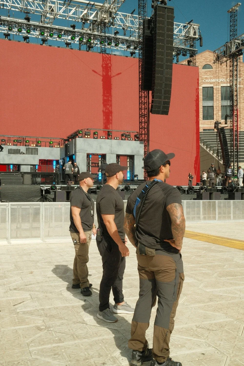 Three security personnel in black shirts and caps stand in a line outside a stage at an event venue, with a large red backdrop and scaffolding and equipment preparing for a performance, under clear blue sky.