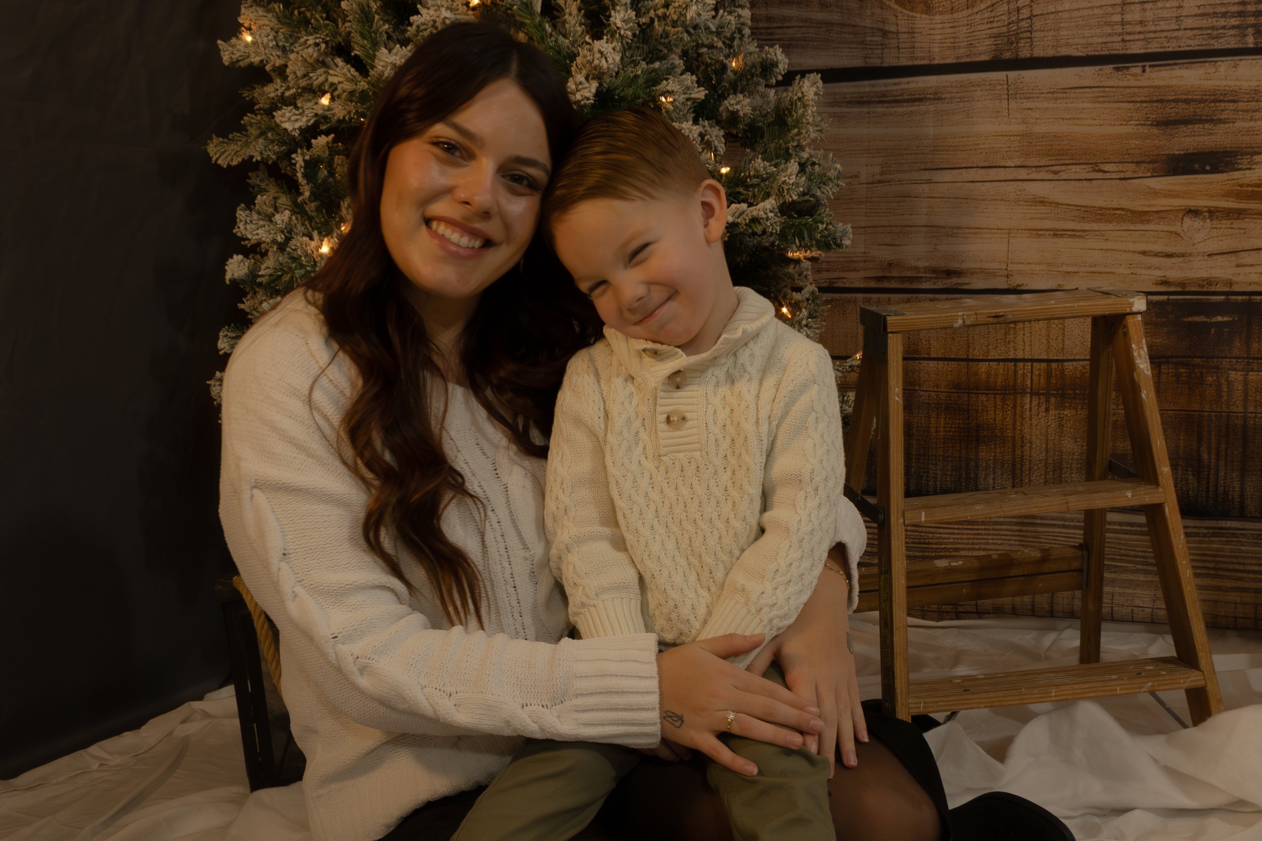 A woman and a young boy with light skin sitting on the floor in front of a decorated Christmas tree, smiling and cuddling together.