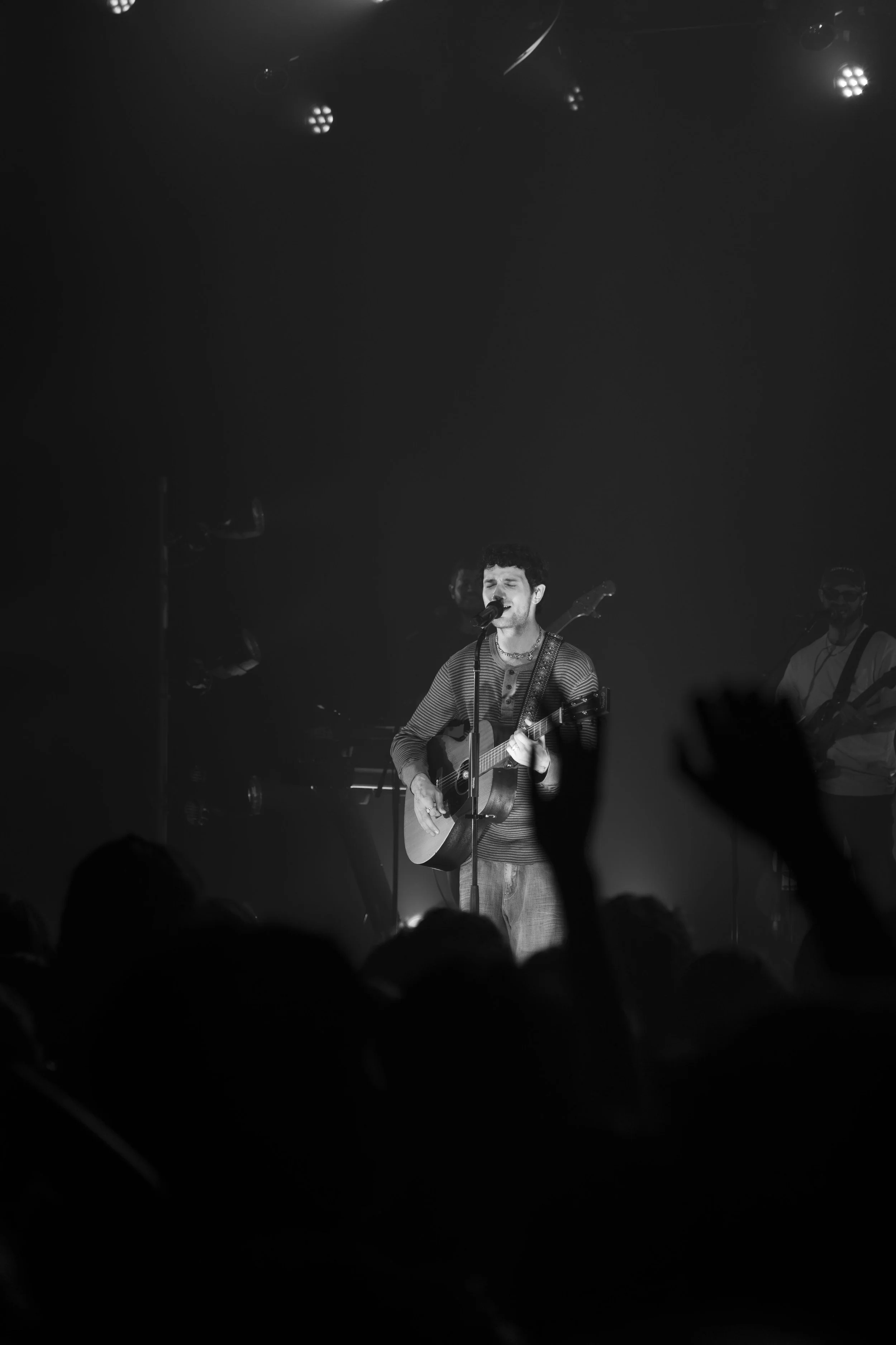 Black and white photo of a male musician performing on stage with an acoustic guitar, singing into a microphone, with backup musicians in the background, and an audience visible at the bottom.