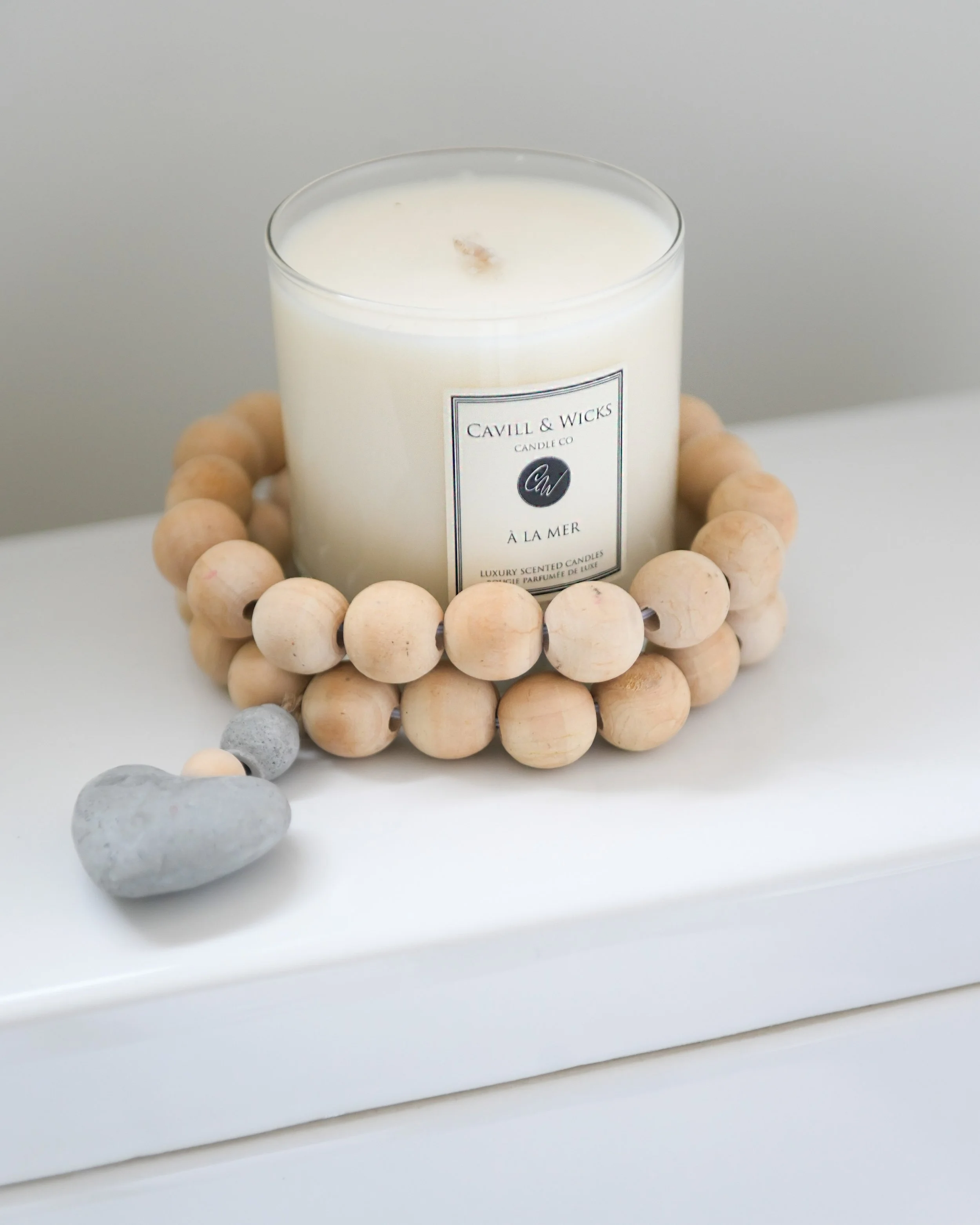 White scented candle in a glass jar with a black and white label, surrounded by a wooden bead ring, placed on a white tray with two small gray stones.
