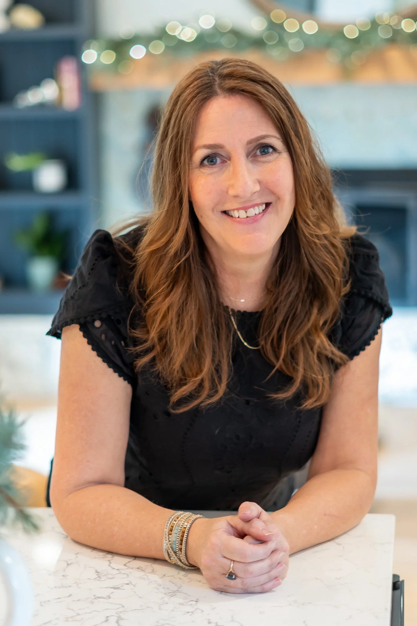 Helen Adams wearing a black top and gold jewelry, sitting at a marble table with her hands clasped, smiling at the camera in a cozy indoor setting.