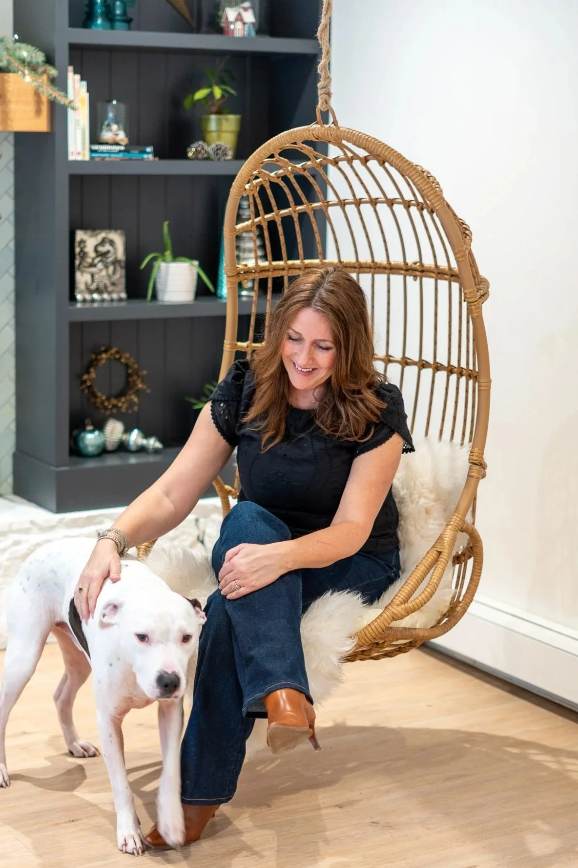 Helen Adams sitting in a hanging wicker chair indoors, petting a white dog.