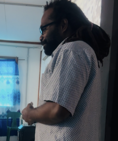 Side view of a person with dark hair and glasses, wearing a white polka-dot shirt, standing indoors near a window with blue curtains