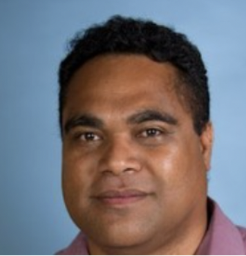 Close-up portrait of a man with dark curly hair and a slight smile against a light blue background.