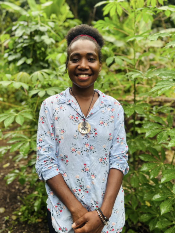 A young woman with dark skin and curly hair tied back with a red headband, smiling, standing outdoors among green plants, wearing a light blue floral shirt and a large necklace with a star-shaped pendant.