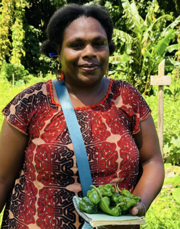 Woman standing outdoors holding a plate of green peppers in a lush, green garden.