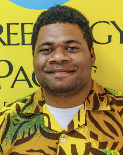 A smiling man with short curly hair wearing a colorful patterned shirt, standing in front of a yellow background with partial black text.