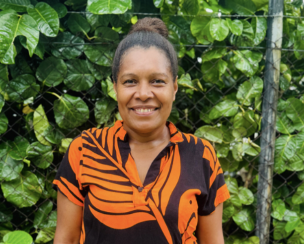 Smiling woman wearing a black and orange patterned shirt, standing outdoors in front of green leafy plants and a chain-link fence.