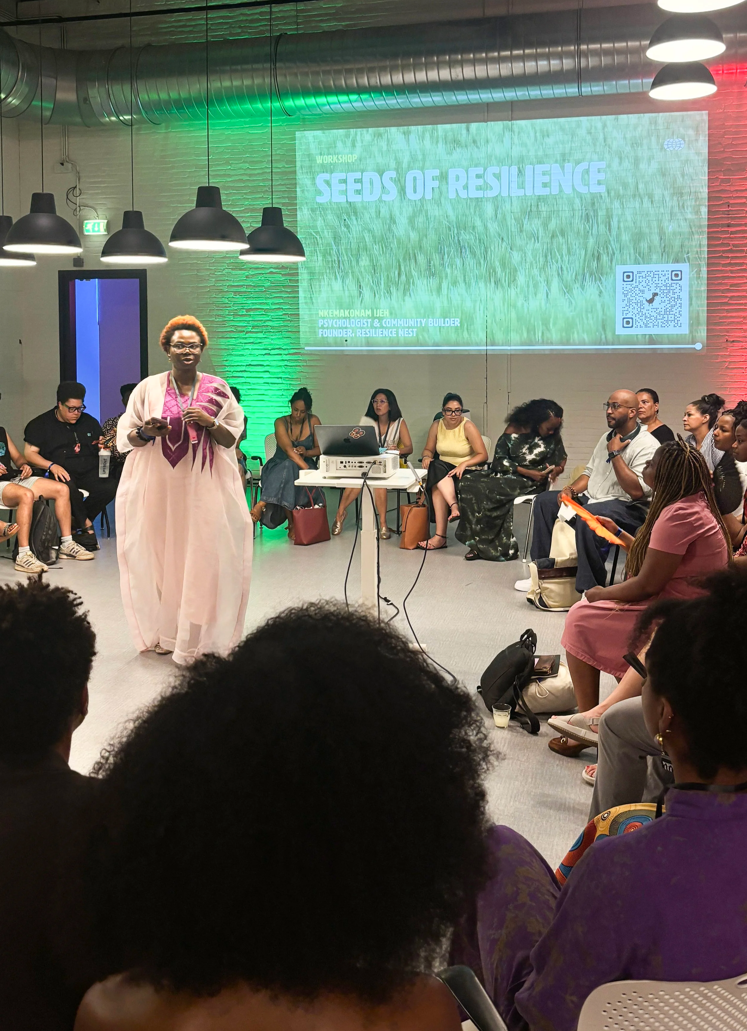 A woman in a pink and white traditional dress standing in front of an audience during a workshop called 'Seeds of Resilience'. The workshop is held in a modern room with a large screen displaying the event's title, and several people are seated, listening attentively.