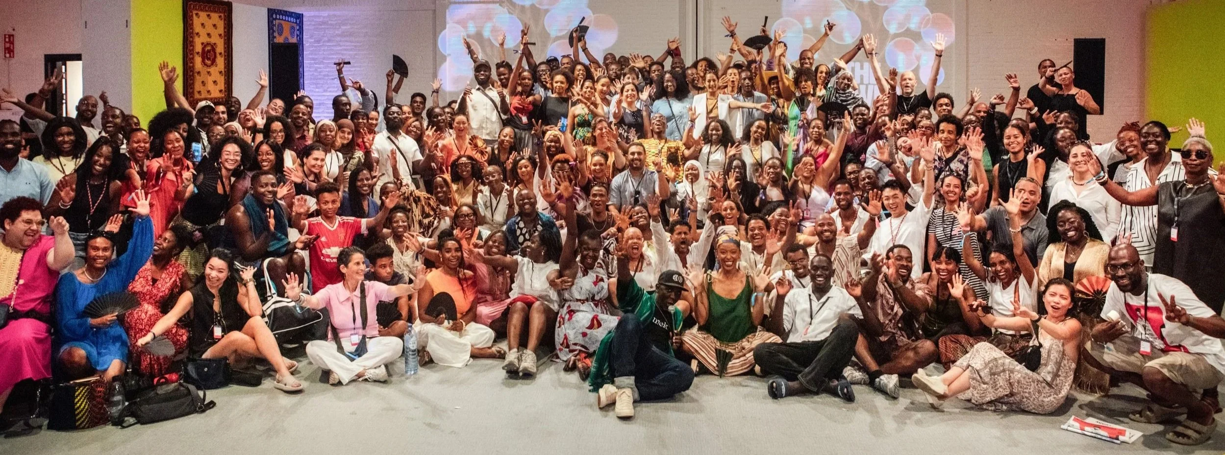 A large group of people gathered indoors, smiling, waving, and celebrating at what appears to be an event or conference.