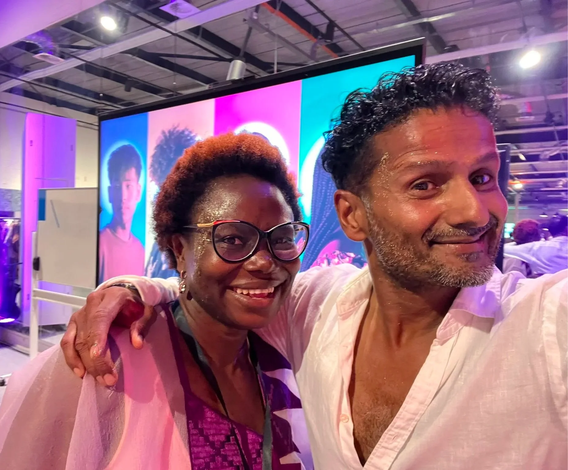 A happy woman with glasses and short reddish hair, hugging a smiling man with dark curly hair, in front of a colorful digital display of a person's portrait with a background of an indoor event space.