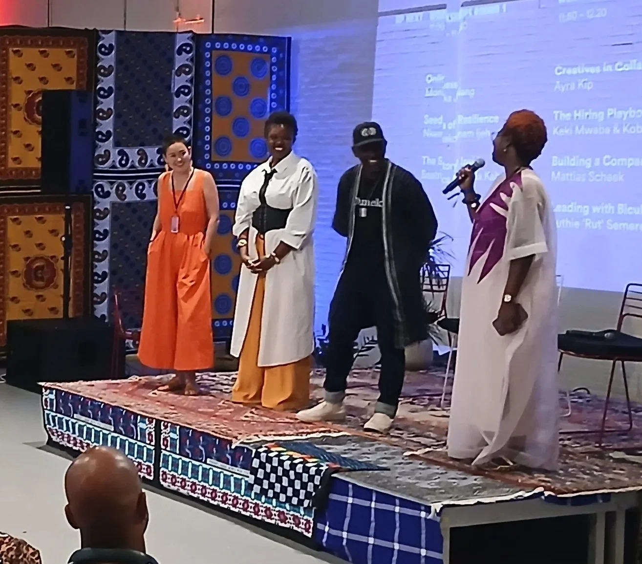 Four people standing on a stage with colorful woven wall hangings and a projection screen behind them. One woman is speaking into a microphone, and the others are listening. They are dressed in vibrant, diverse clothing.