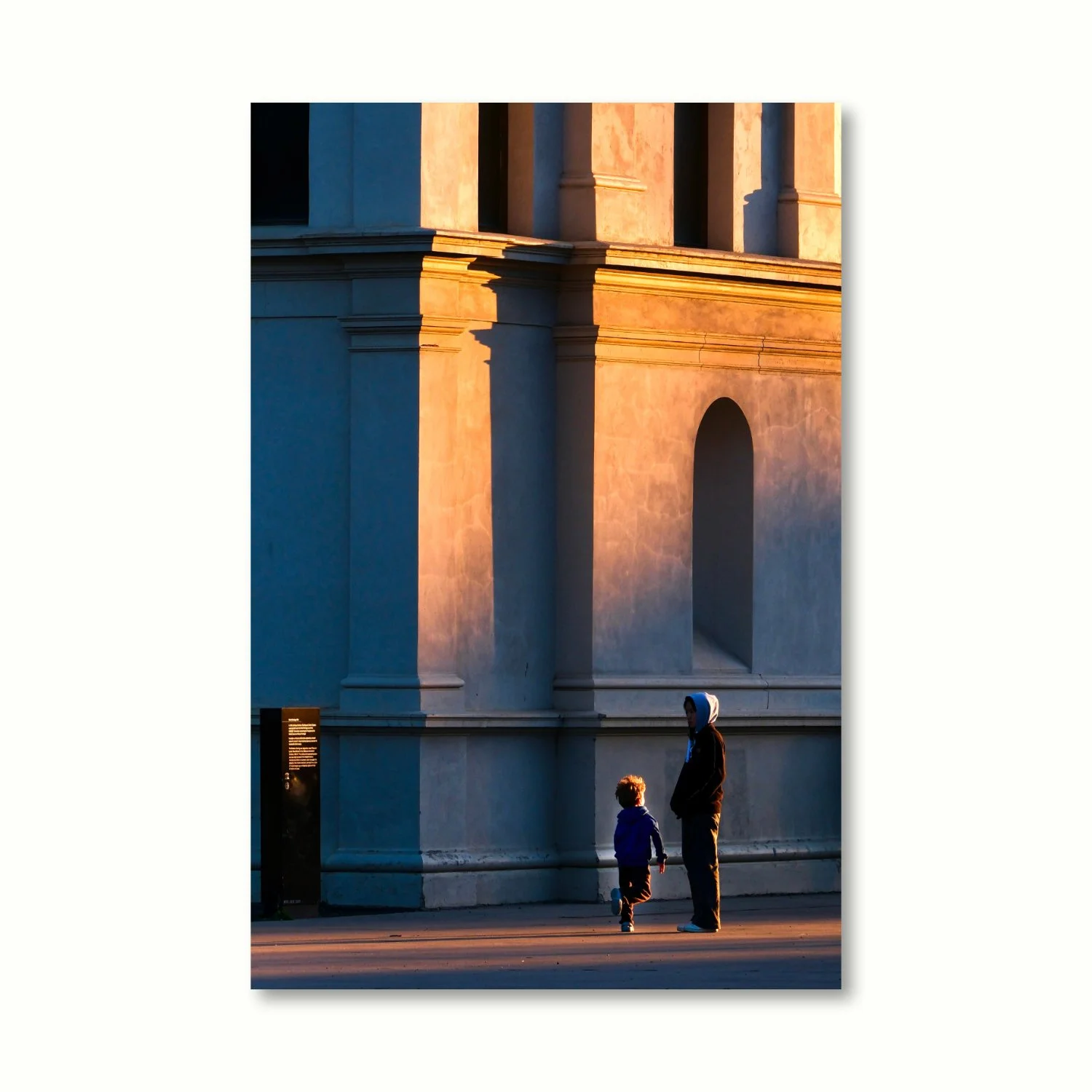 A mother and child pause near a golden-lit classical building, their figures partially caught in the warm evening light & long shadows.