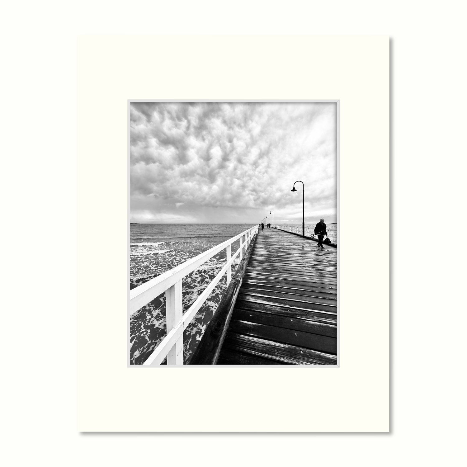 A frame mount containing a B&W print of dramatic storm clouds rolling in over the pier with a lone fisherman leaving.