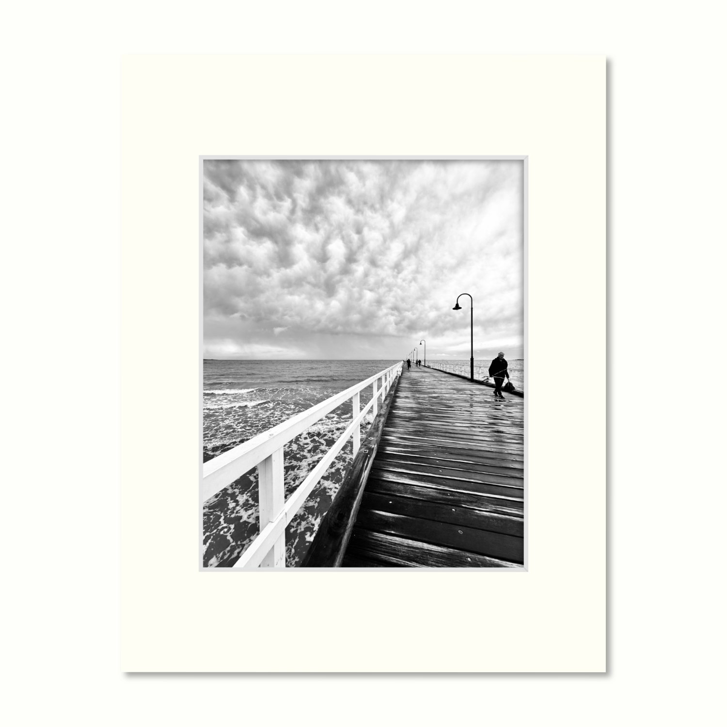 A frame mount containing a B&W print of dramatic storm clouds rolling in over the pier with a lone fisherman.
