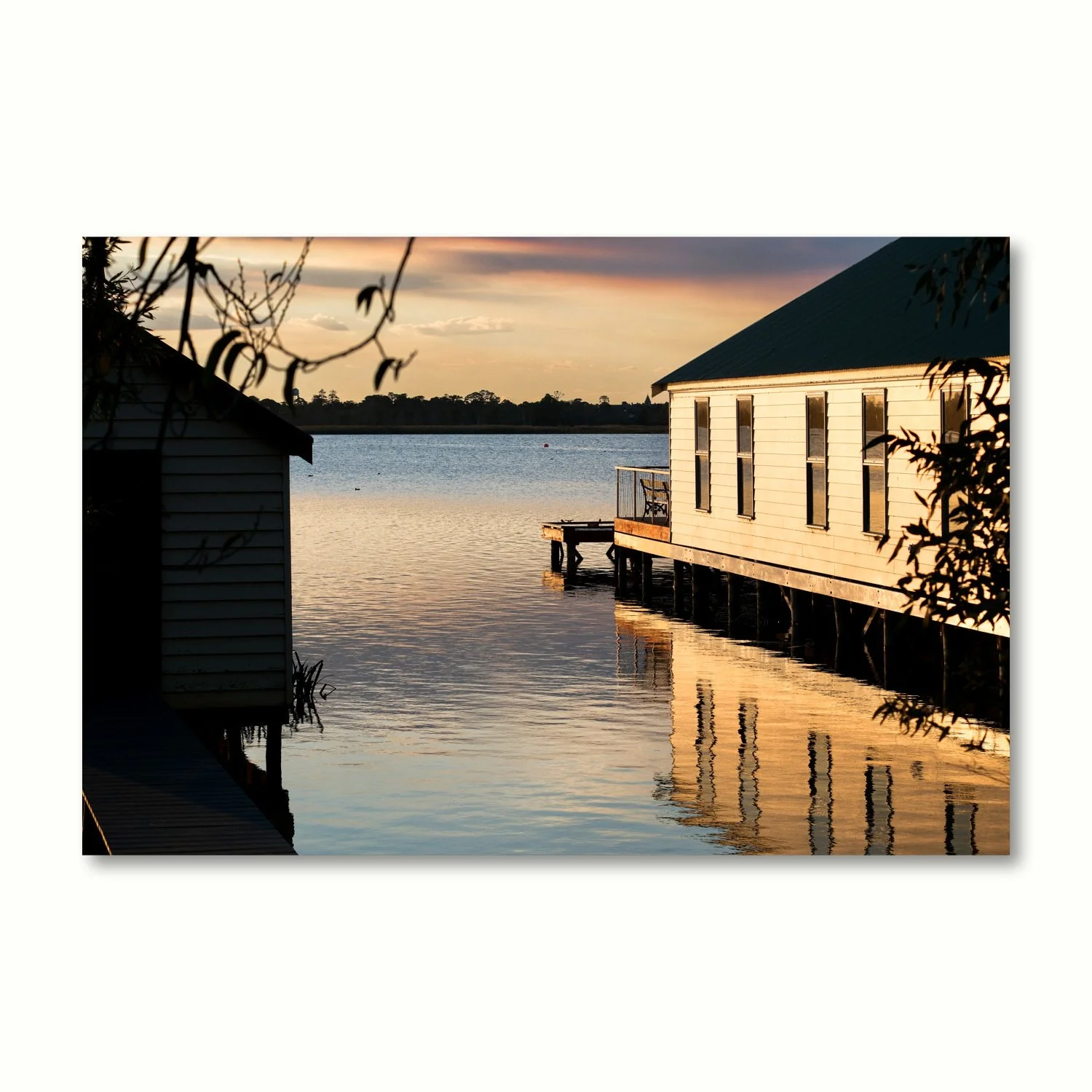 A single print of boatsheds on a lake at sunset, with golden water reflections.