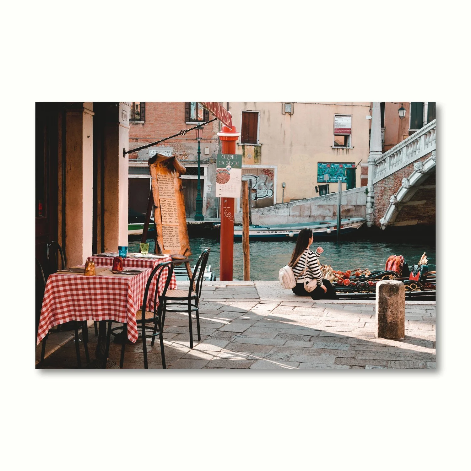A peaceful scene of a girl waiting patiently by the canal and adjacent to a charming trattoria with outside tables set for dinner.