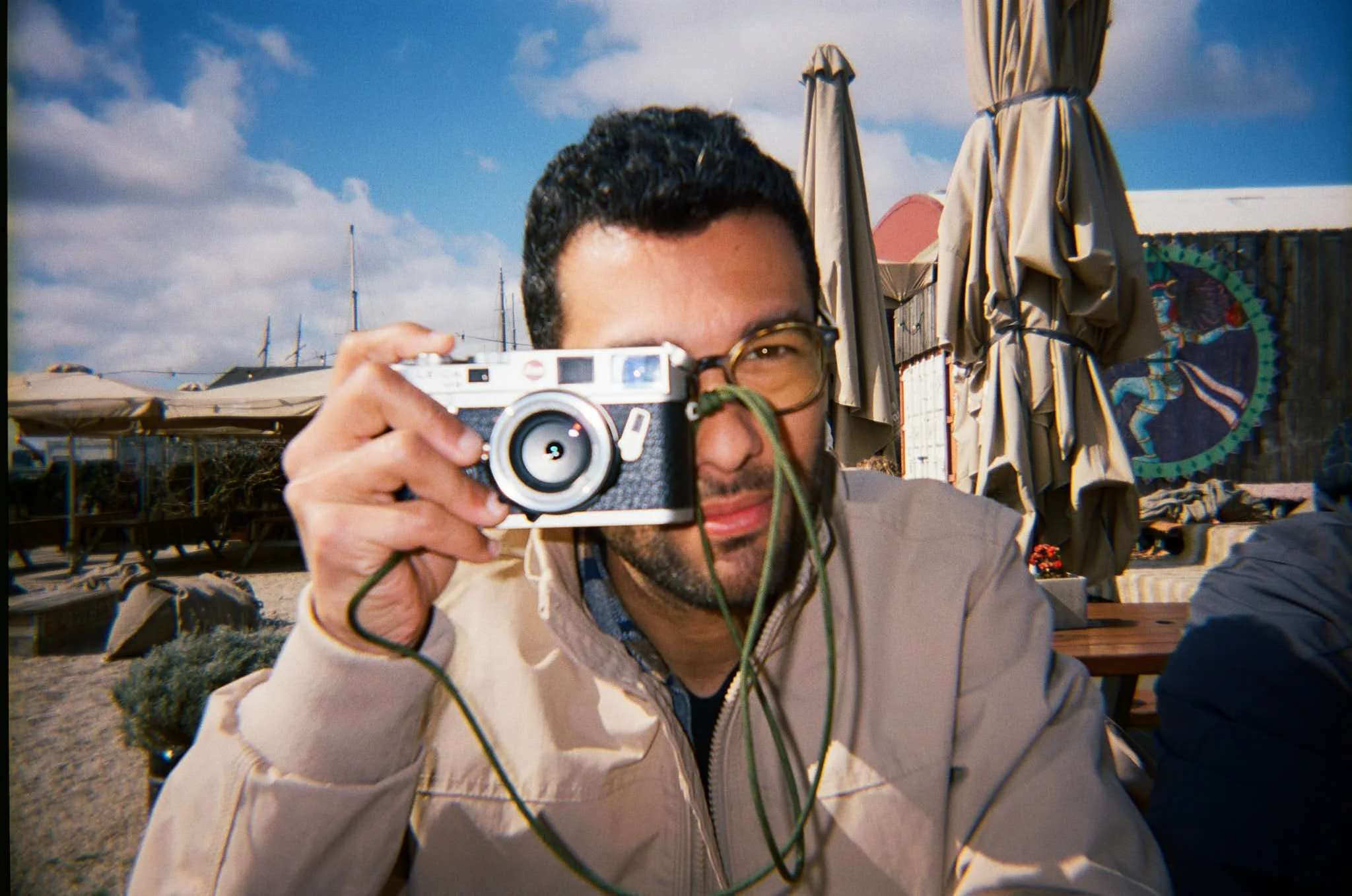 Man with glasses taking a photo with a vintage camera at an outdoor setting, with umbrellas and a painted wall in the background.