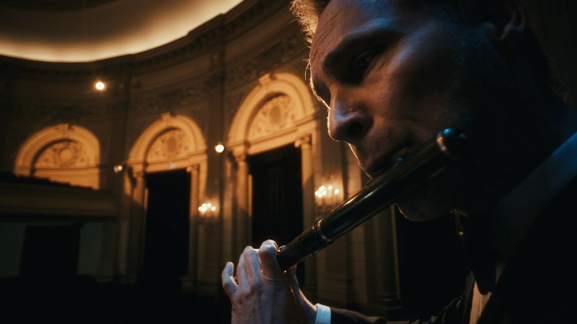 A man in a tuxedo playing a flute in an ornate, dimly lit room with classical architectural details.