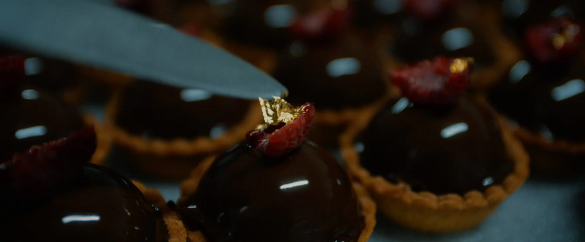 Close-up of small tartlets topped with glossy chocolate ganache and a red berry, with a spoon adding a topping.