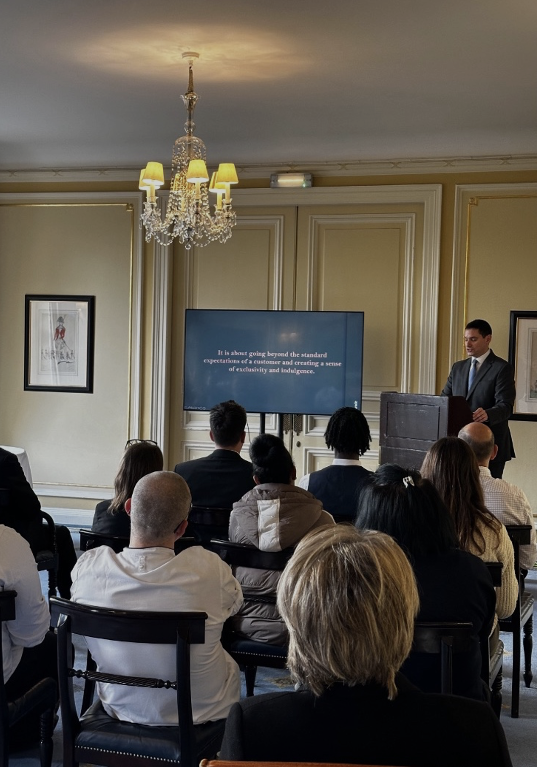 A man giving a presentation in a formal conference room with an audience seated facing him. A large screen displays text about going beyond standard expectations of a customer, located on ornate walls with artwork and a chandelier overhead.