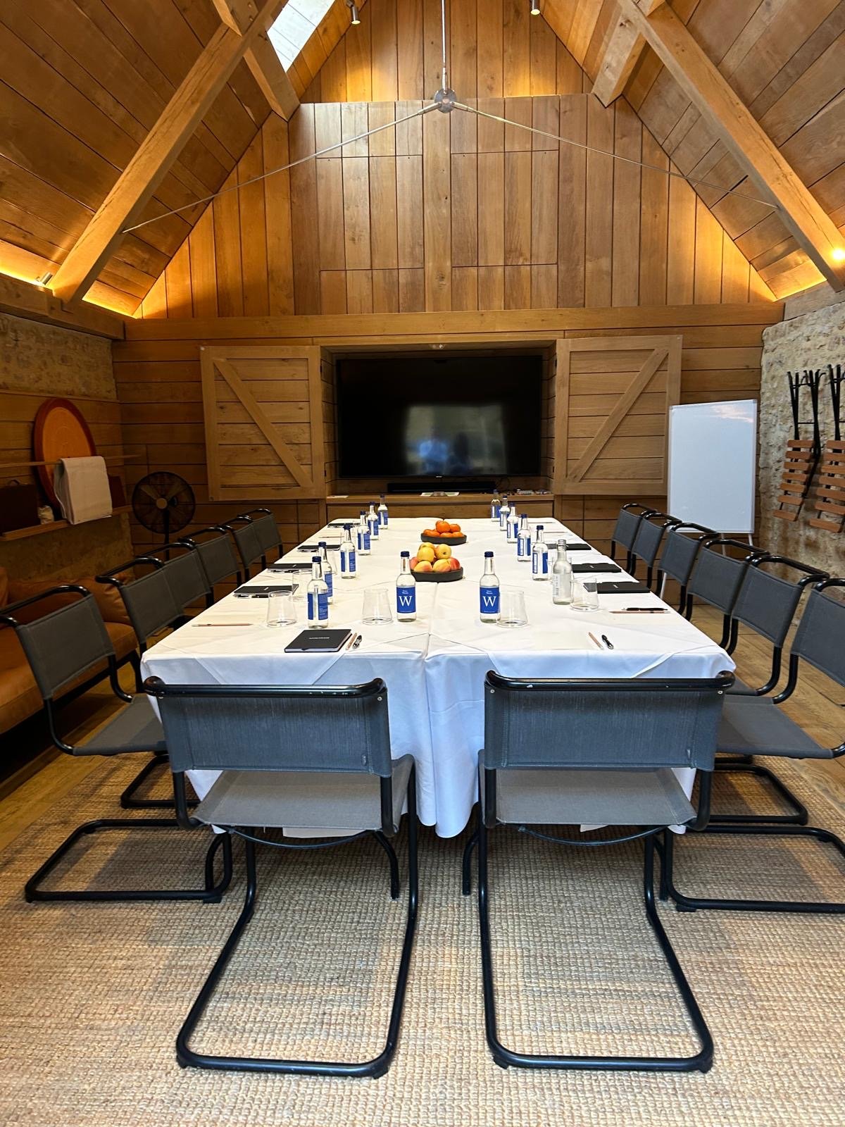 Conference room with a long rectangular table covered with a white tablecloth, set with water bottles, glasses, notepads, and pens. The room has wood-paneled walls and ceiling, with a large flat-screen TV at the front, a whiteboard, and a stone wall 