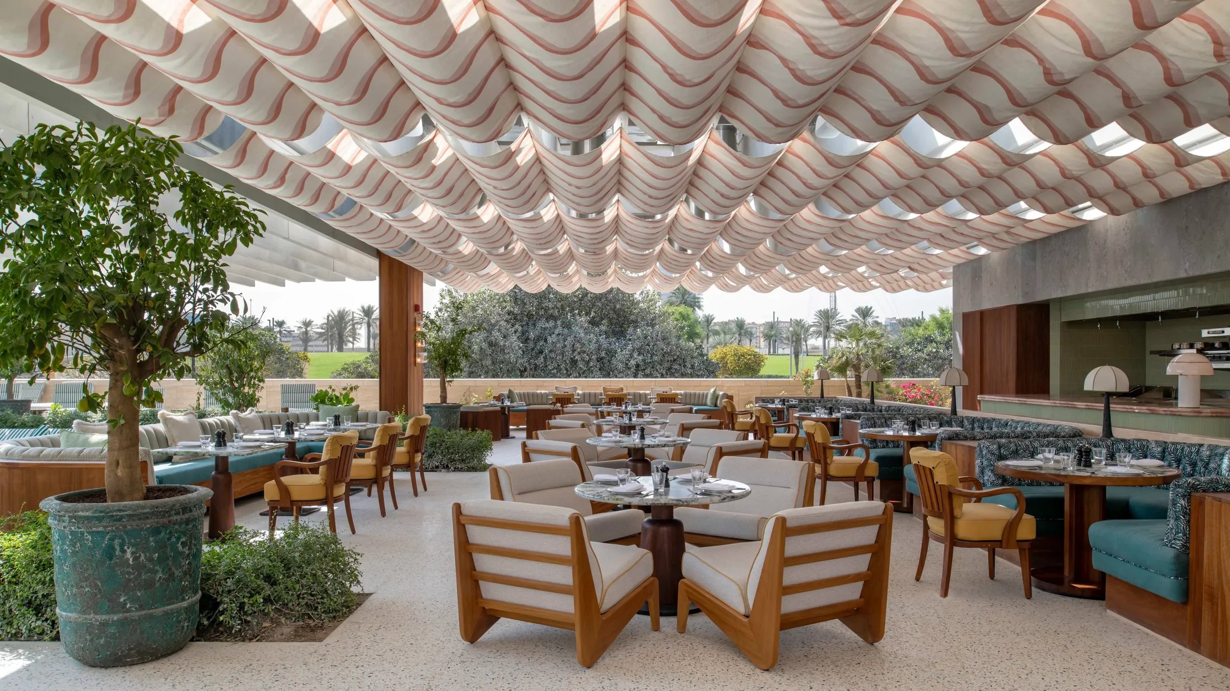 Open-air restaurant with round tables, cushioned chairs, potted trees, and a striped fabric canopy overhead, overlooking a grassy area with palm trees.