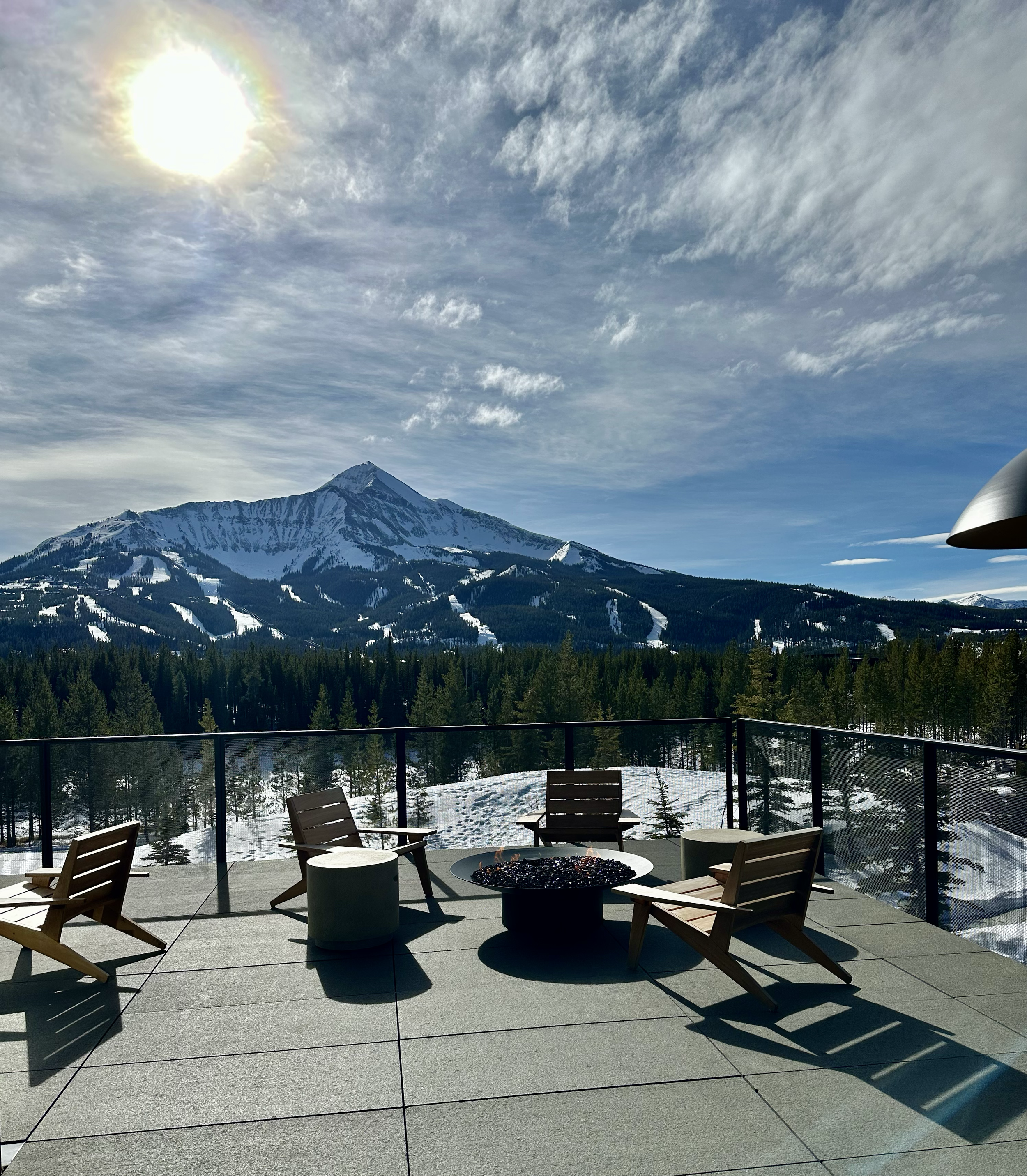 A mountain landscape with snow-capped peaks and a partly cloudy sky, viewed from an outdoor patio with chairs, tables, and a firepit.