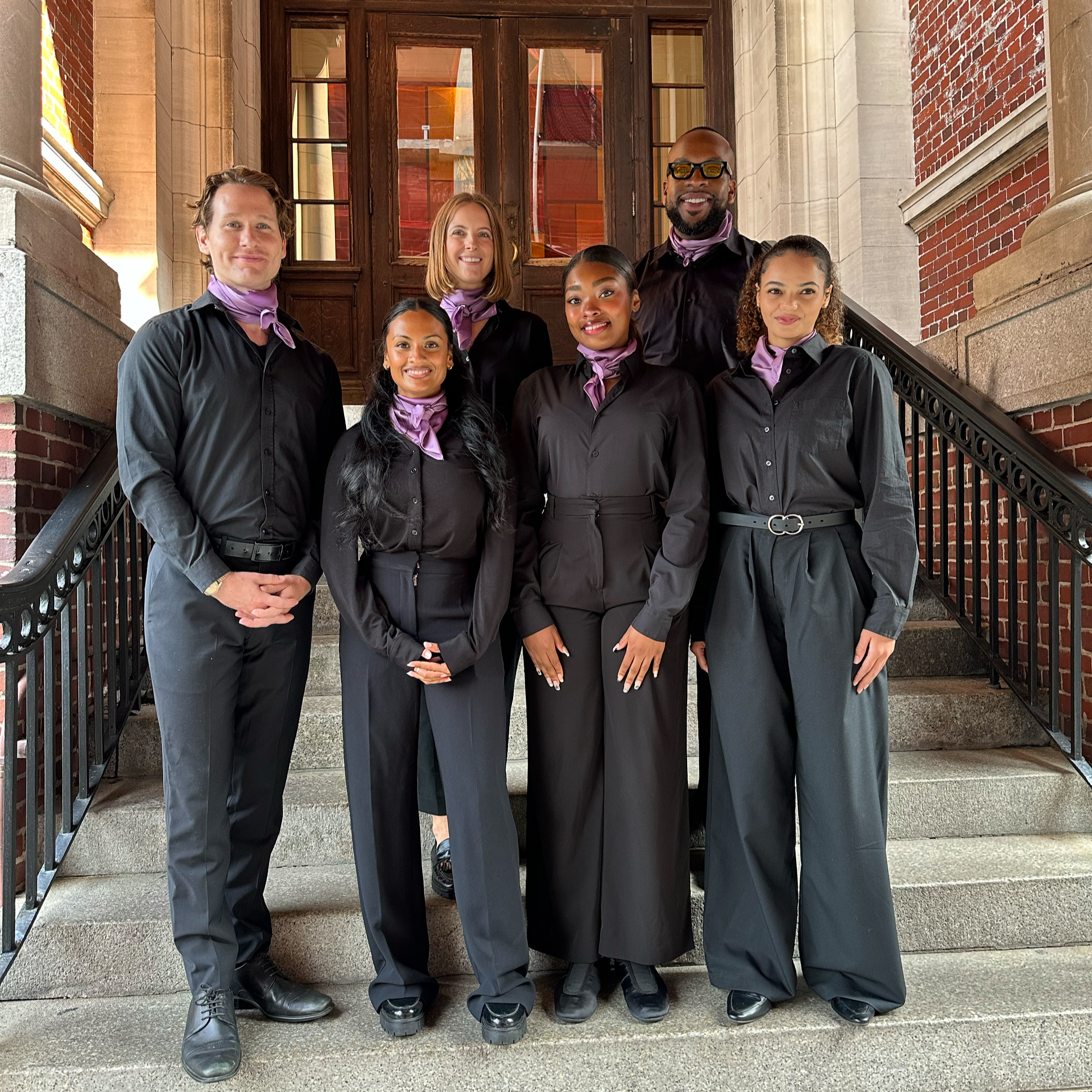 A diverse group of seven people dressed in black with purple scarves stands on a staircase outside a building with brick and stone walls and large wooden doors. They are smiling and looking at the camera.