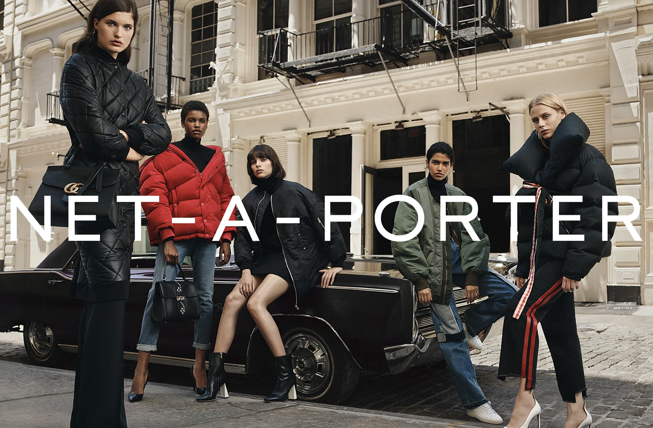 A group of five diverse young women in fashionable winter outfits stands on a city street in front of a vintage car and a multi-story building with fire escapes, posing confidently for a fashion photoshoot.