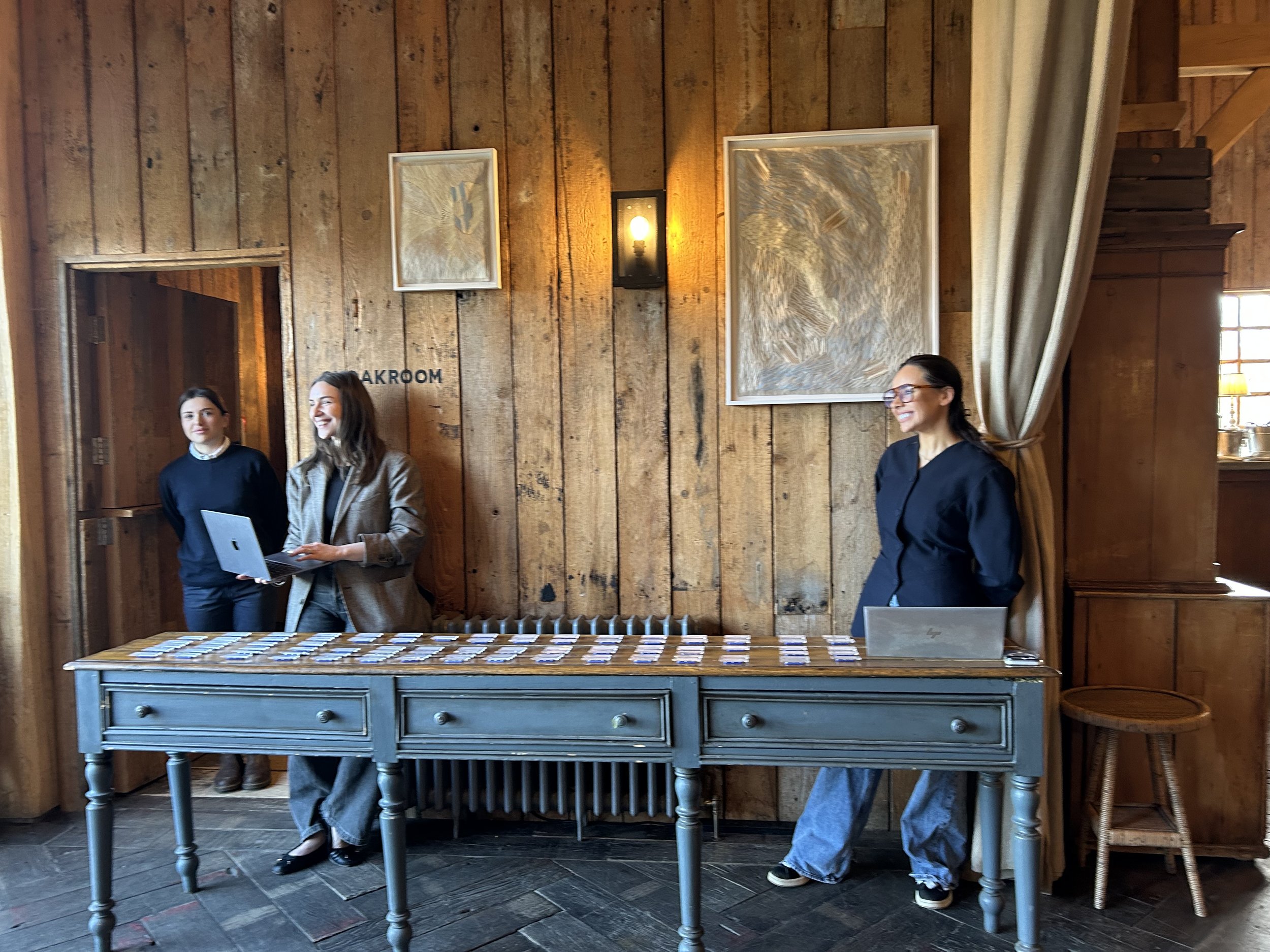 Three women standing behind a table with cards on it in a room with wooden walls and framed art, one woman holding a laptop, the others smiling.