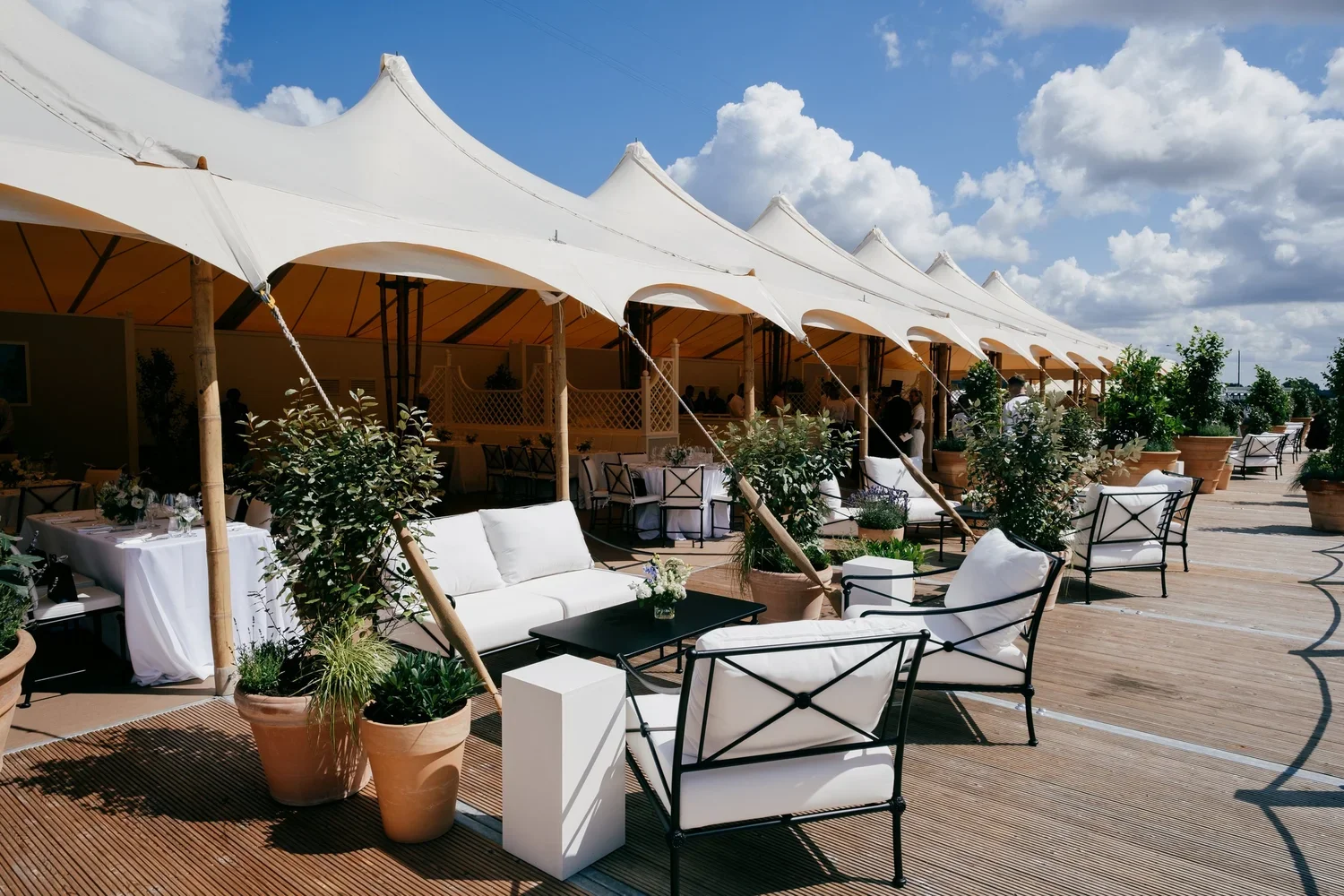 Outdoor patio with white canopy tents, white cushioned chairs, black tables, and potted plants under a partly cloudy sky.