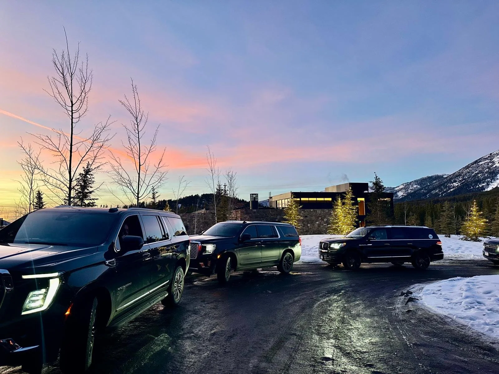 A parking lot with several black SUVs parked on a snowy surface, with snow-covered mountains and a colorful sky at sunset in the background.