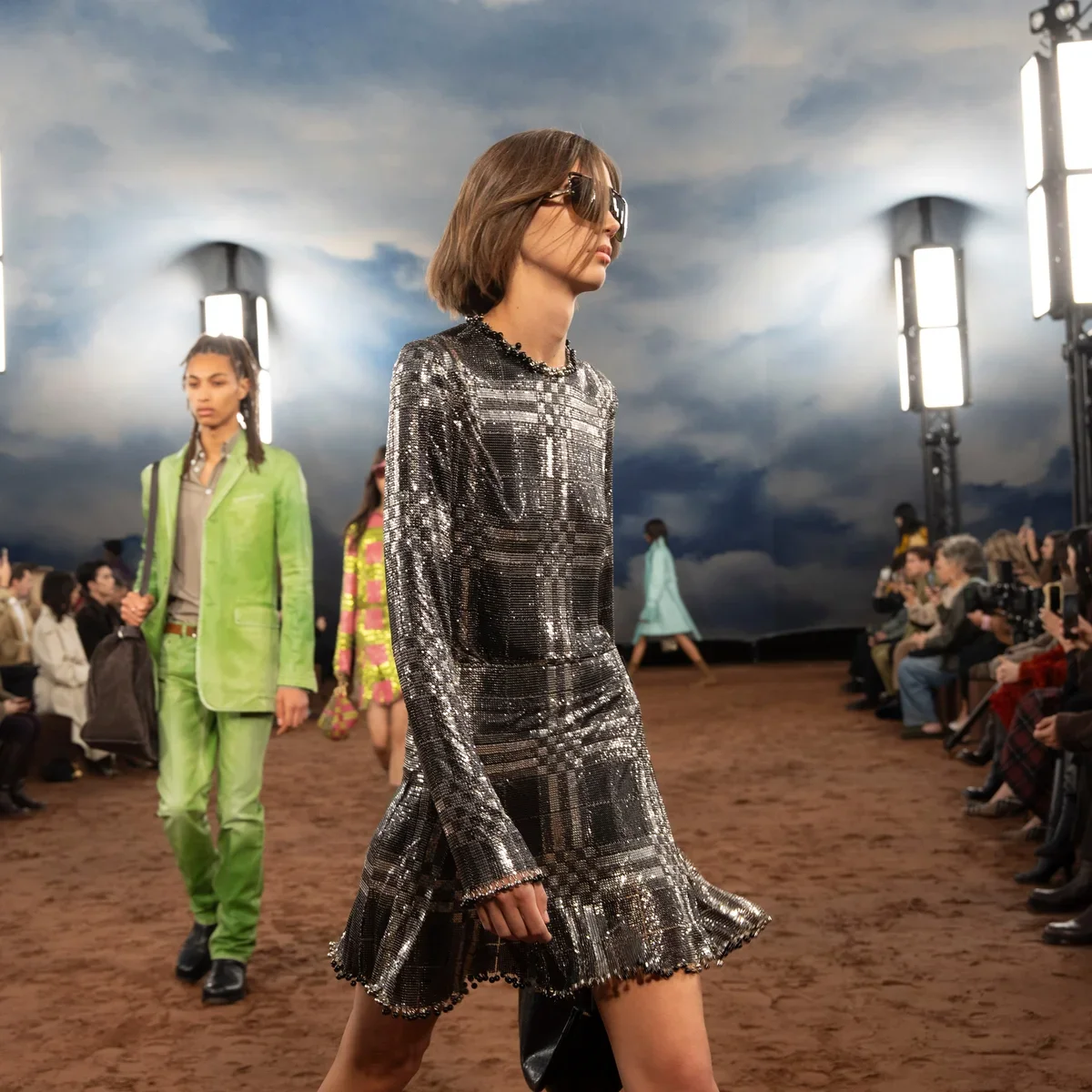 Models runway walking during a fashion show with a cloudy sky backdrop, audience seated on both sides, and large studio lights.