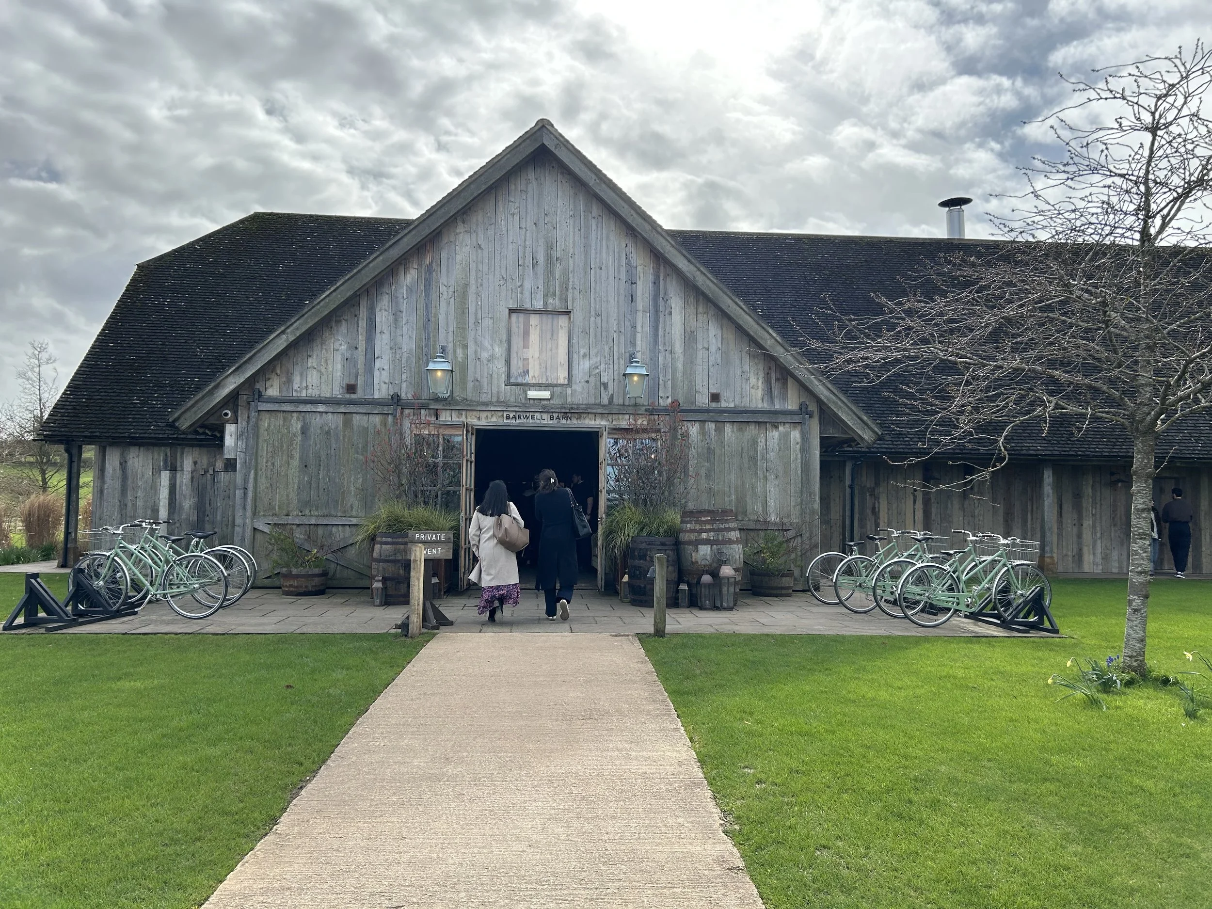 People walking towards a rustic barn with a sign that reads 'Barwell Barn,' surrounded by bicycles and green grass, under a cloudy sky.