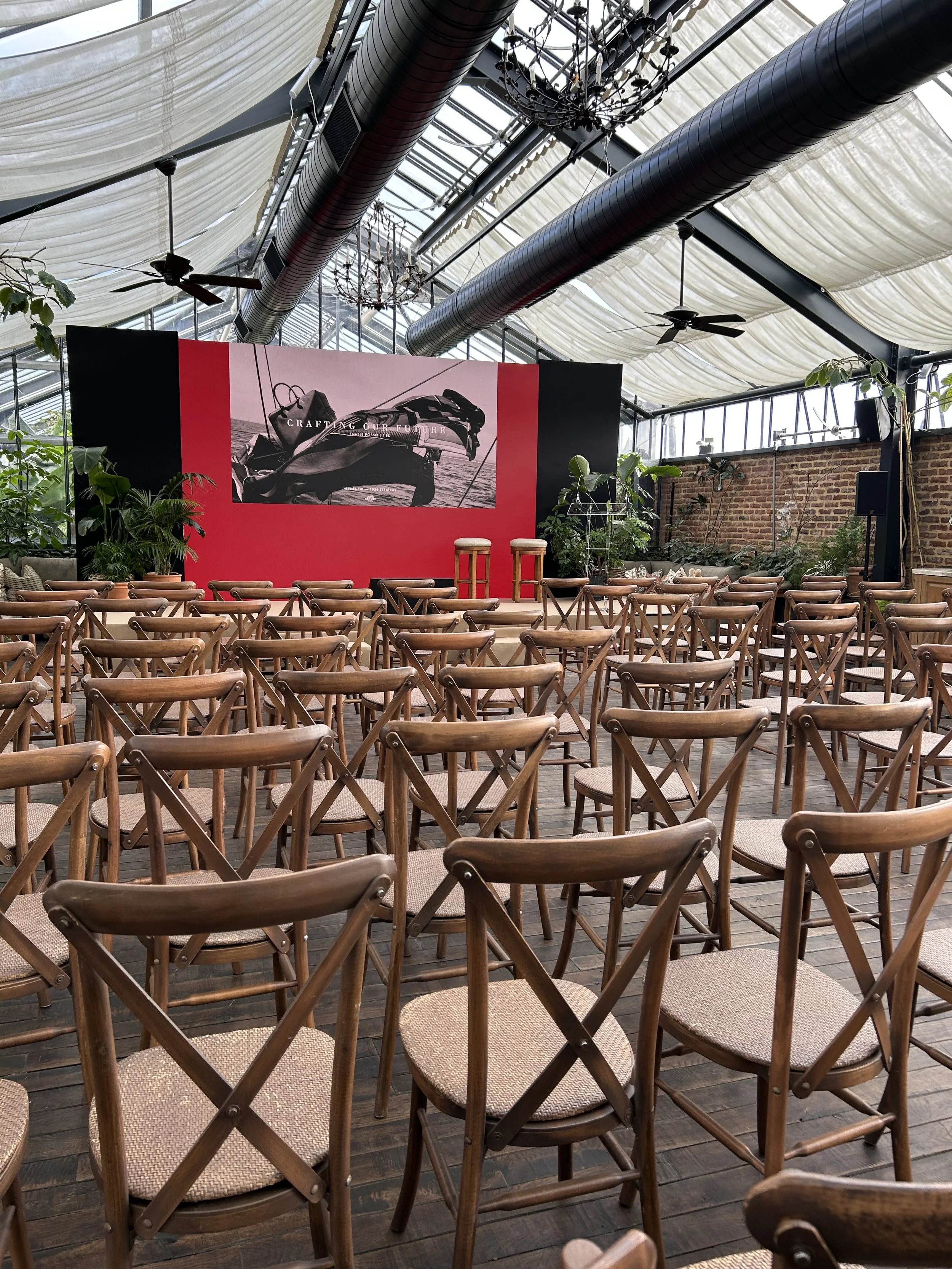 A large indoor event space with wooden chairs arranged facing a stage with a red backdrop and a black and white banner. There are plants along the sides, ceiling fans, exposed ductwork, and chandeliers hanging from the ceiling made of fabric and glas