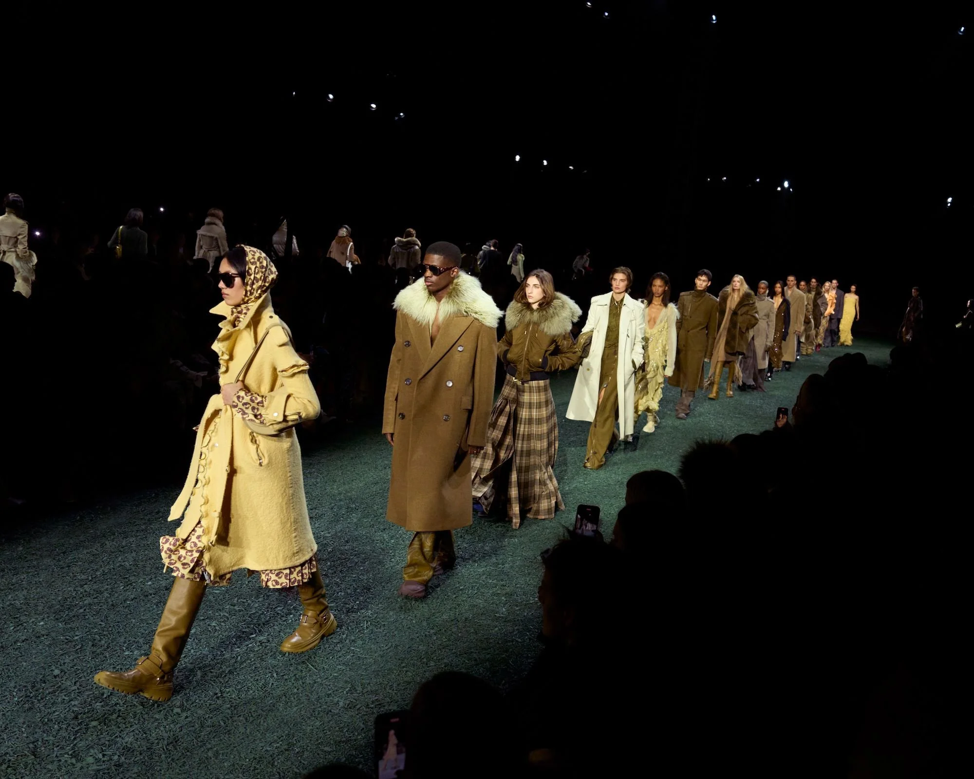 Models walking on a runway during a fashion show with dark background.