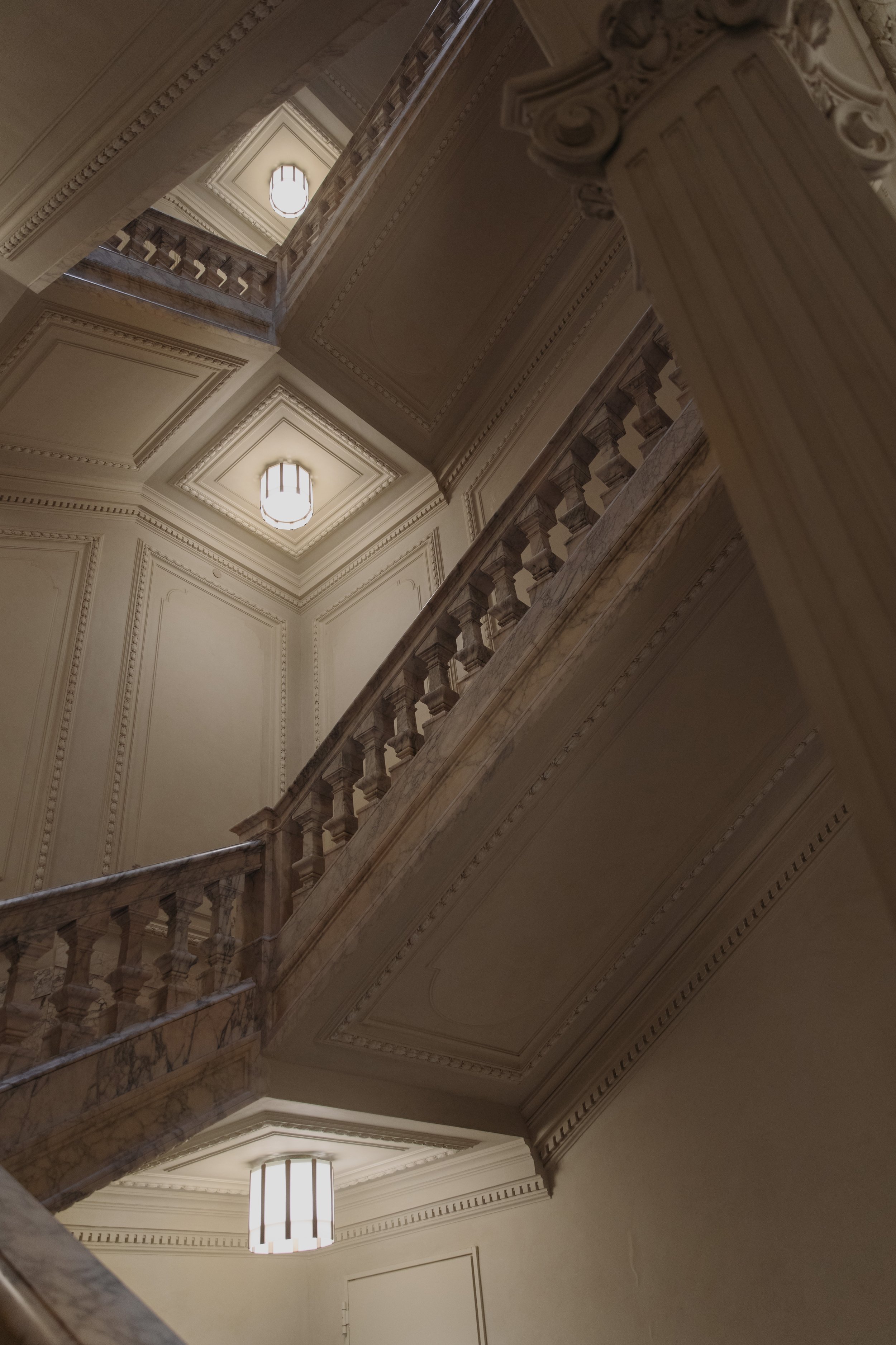 Interior view of a grand staircase and ornate ceiling with two lights, shot from below.