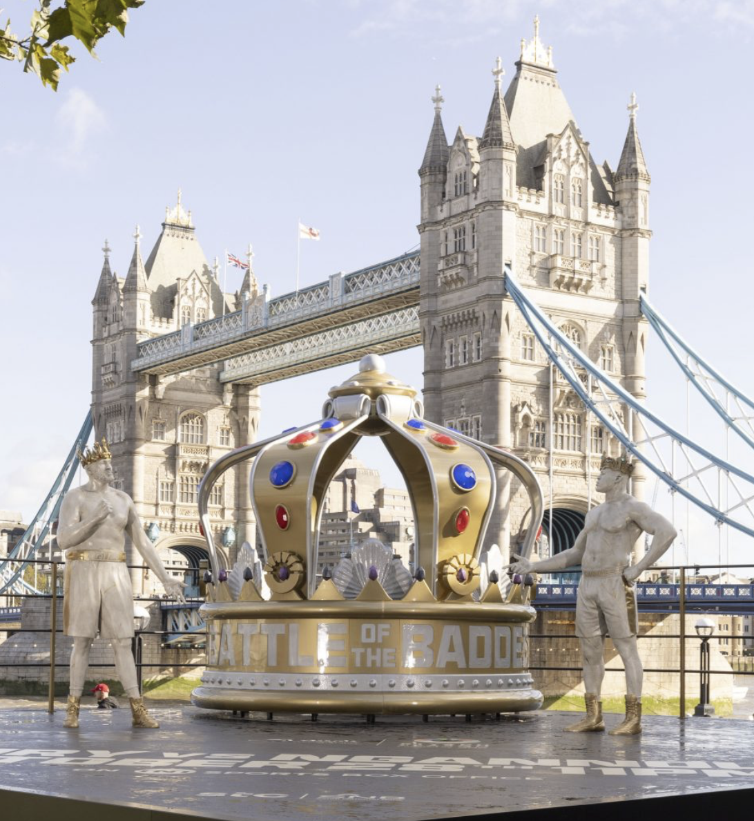 A decorative stage with a crown prop labeled 'Battle of the Badge' in front of London's Tower Bridge, flanked by statues of two boxers.